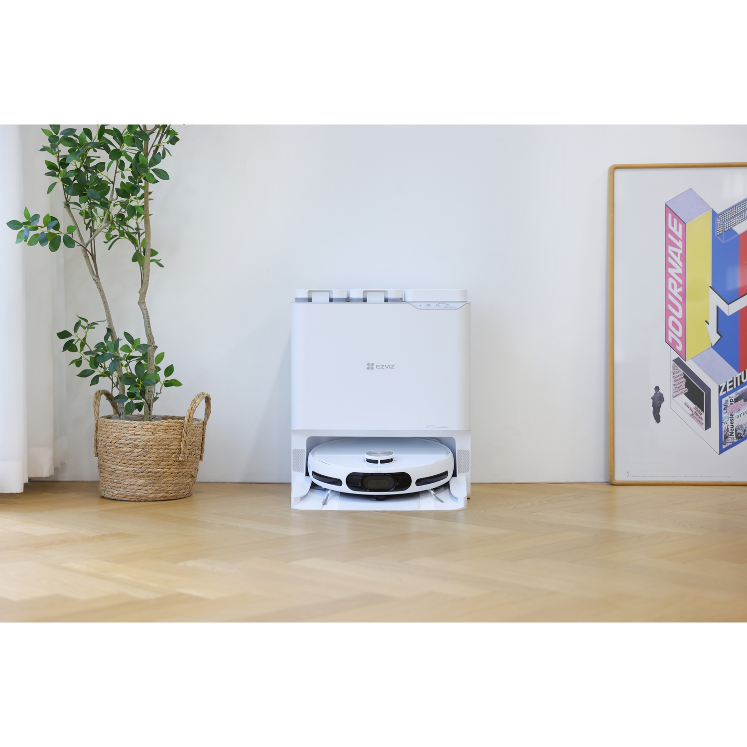 Een witte robotstofzuiger staat in zijn oplaadstation. Een plant in een pot en een ingelijste poster staan aan de zijkanten.