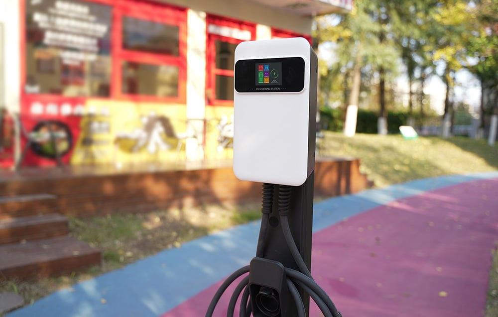 Egy elektromosautó-töltőállomás egy elmosódott épület előtt áll. Az állomás fehér és fekete, töltőkábelekkel.