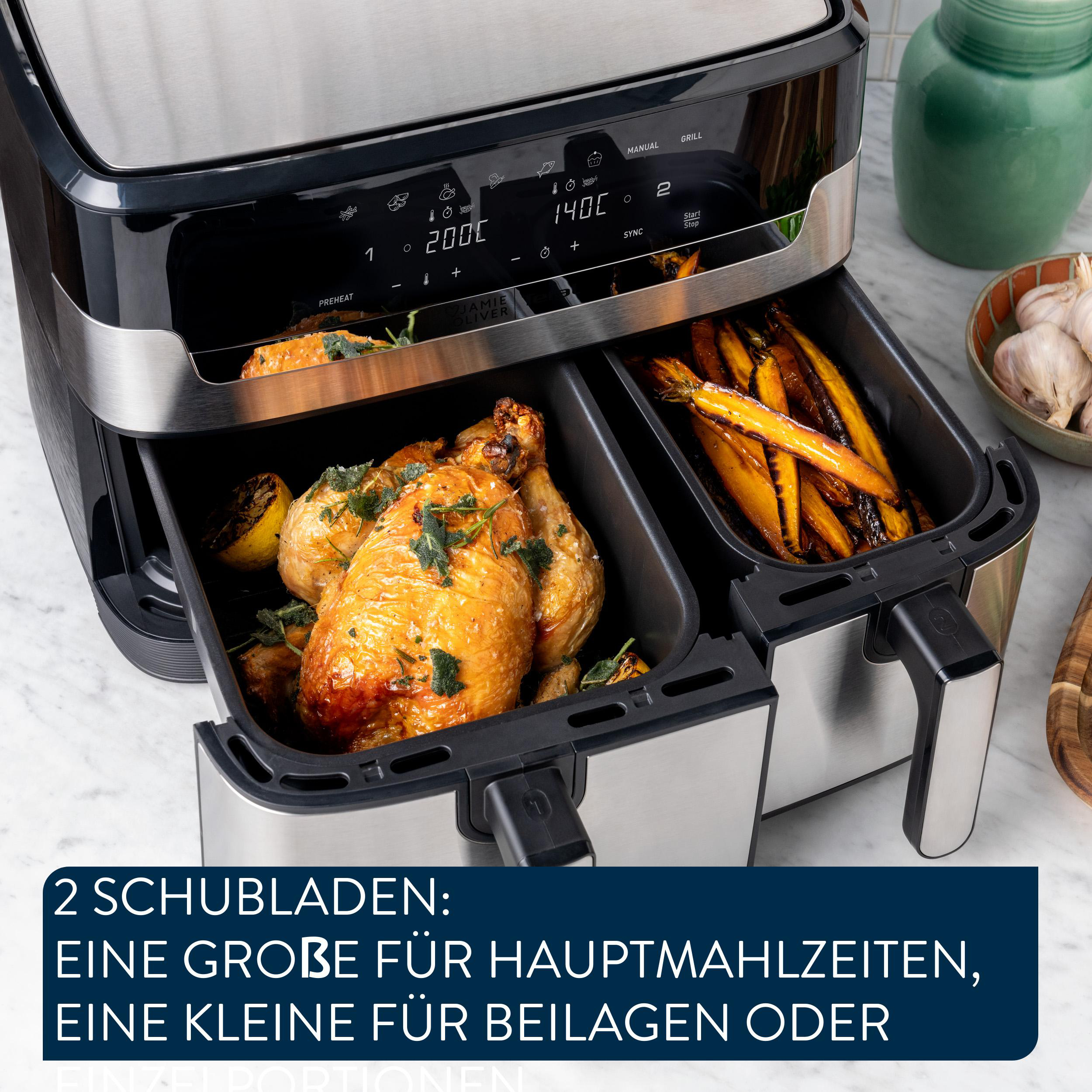 Eine Heißluftfritteuse mit zwei Fächern, eines mit Brathähnchen, das andere mit Karotten. Eine Schüssel mit Knoblauch ist sichtbar.