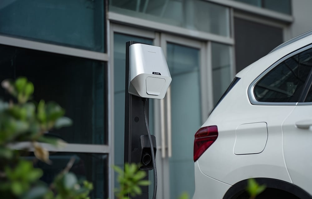 Egy elektromos autó töltődik. Egy fehér autó parkol egy töltőállomás mellett. Zöld lombozat van az előtérben.