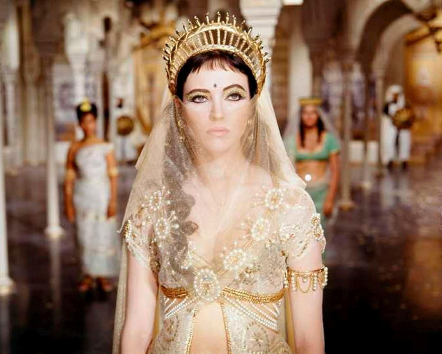 Frau mit goldenem Diadem und Gewand. Andere im Hintergrund. Szene ist ein Palast oder Tempel mit Säulen und Bögen.