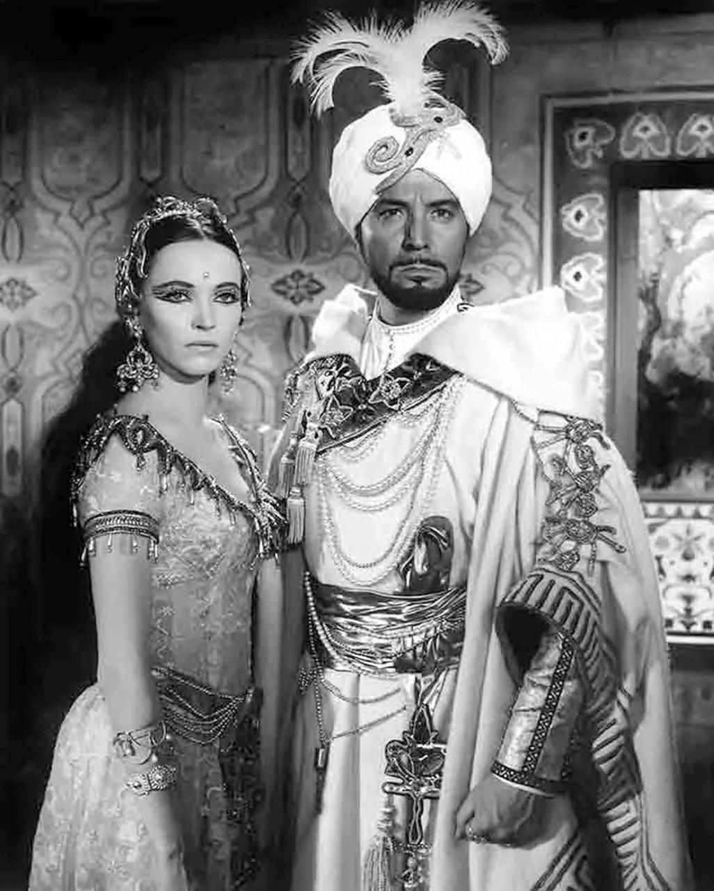 Ein Mann und eine Frau in aufwändiger Kleidung stehen nebeneinander. Der Mann trägt einen Turban und einen Umhang, die Frau ein Juwelenkleid.