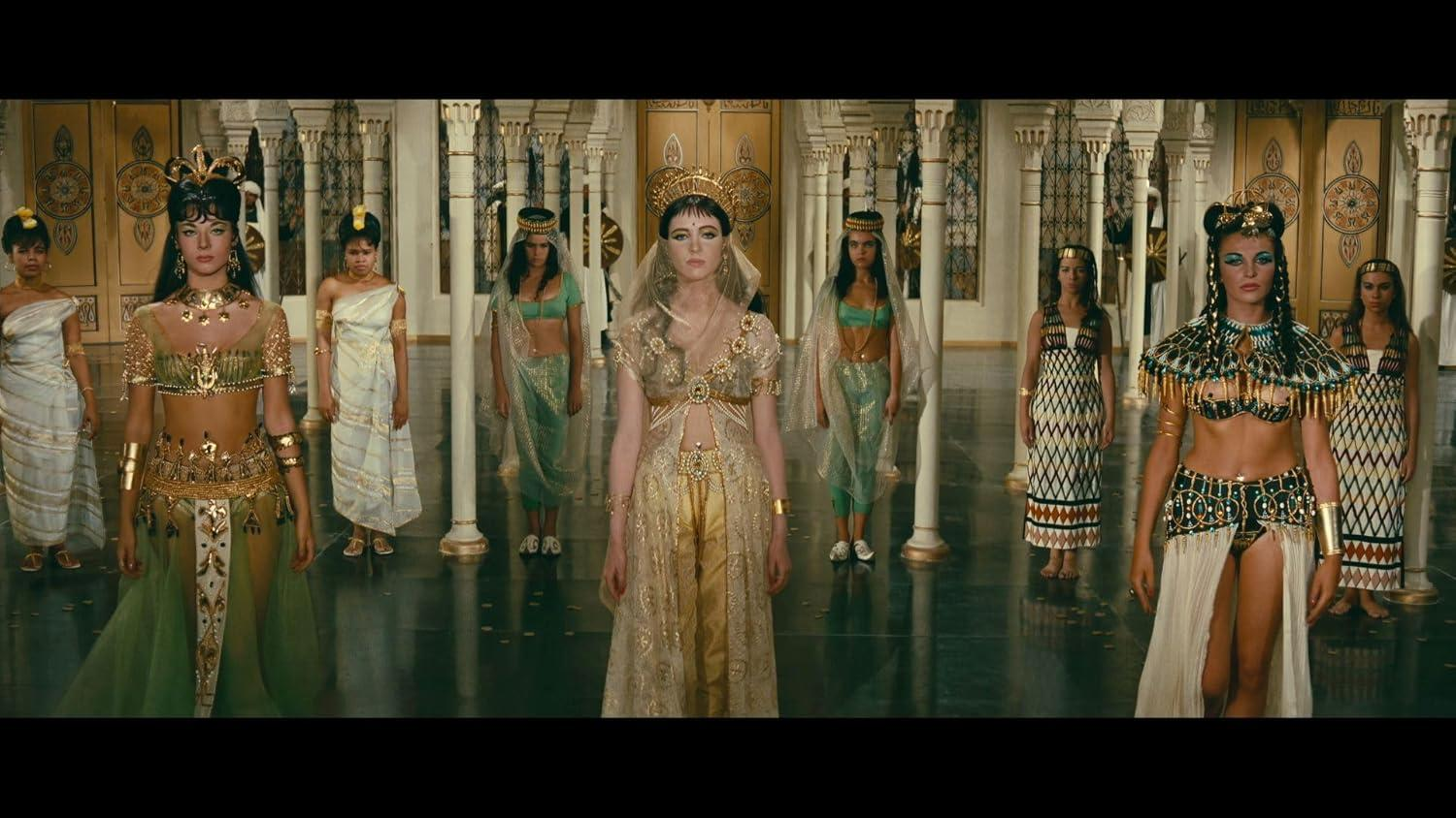 Eine Gruppe von Frauen in altägyptischer Kleidung steht in einem Palast. Gold- und Grüntöne dominieren die Szene, im Hintergrund Säulen.