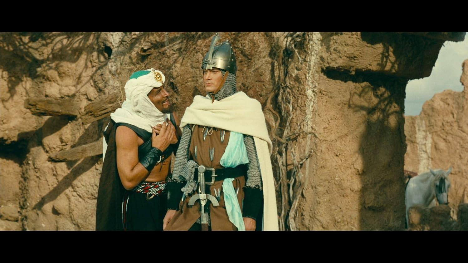 Zwei Männer in historischen Kostümen stehen in einer Wüstenlandschaft. Einer in Rüstung, der andere in traditioneller arabischer Kleidung. Ein weißes Pferd ist in der Nähe.