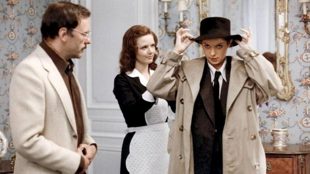 Eine Person in Trenchcoat und Hut passt den Hut an. Zwei andere beobachten. Blumige Tapete im Hintergrund.