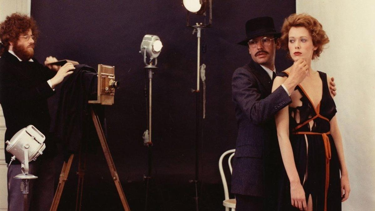 Ein Fotograf mit einer Großformatkamera, zwei Männer und eine posierende Frau. Die Szene spielt in einem Studio mit Beleuchtung.