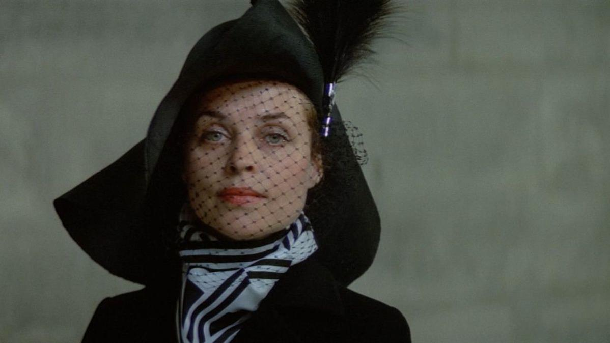Eine Frau mit schwarzem Hut, Schleier und Schal. Sie hat roten Lippenstift und steht vor grauem Hintergrund.