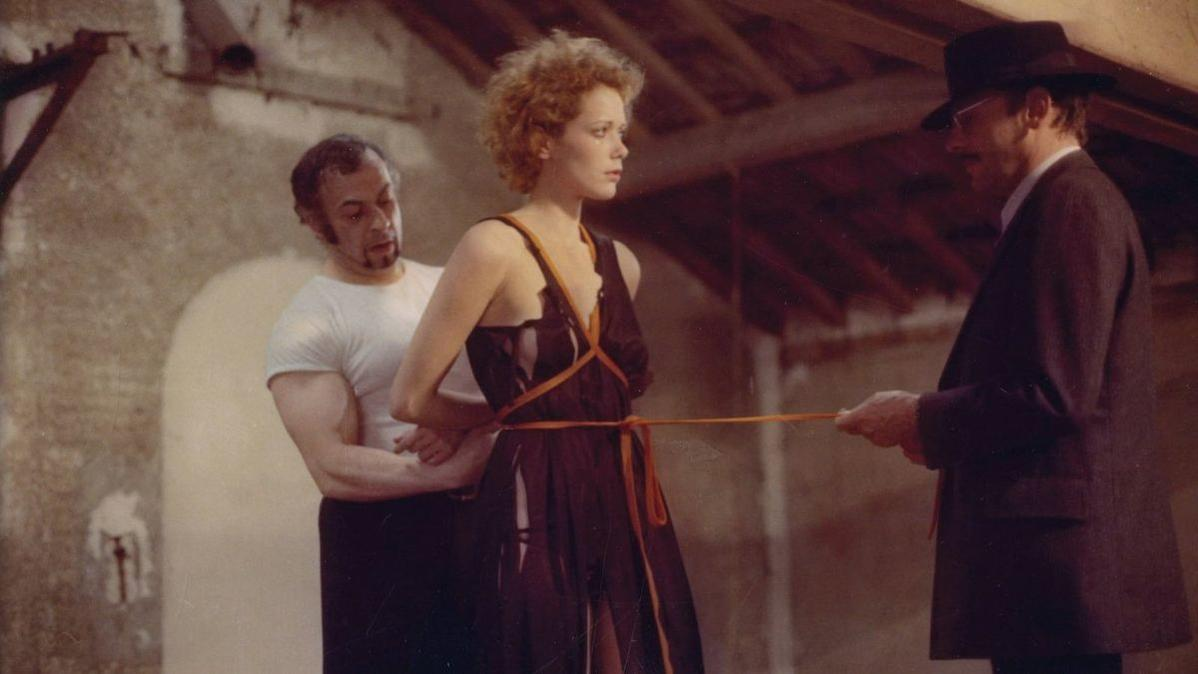 Eine Frau in einem dunklen Kleid ist mit einem Seil gefesselt. Zwei Männer sind anwesend; einer hinter ihr und ein anderer hält das Seil. Szene in einem Gebäude.