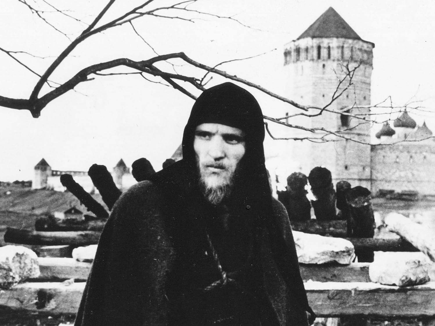 Ein Mann in einem Kapuzenmantel steht vor einer Festung. Kahle Äste rahmen den Himmel ein. Der Mann hat einen Bart und schaut in die Kamera.