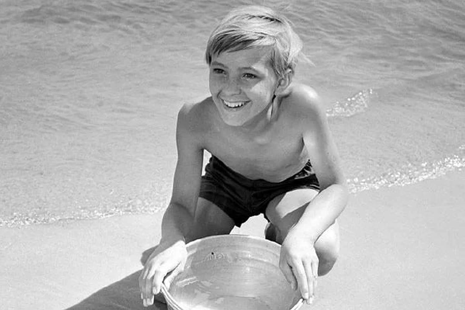 Ein lächelnder Junge kniet am Strand und hält einen Eimer. Das Meer ist im Hintergrund. Das Bild ist schwarzweiß.