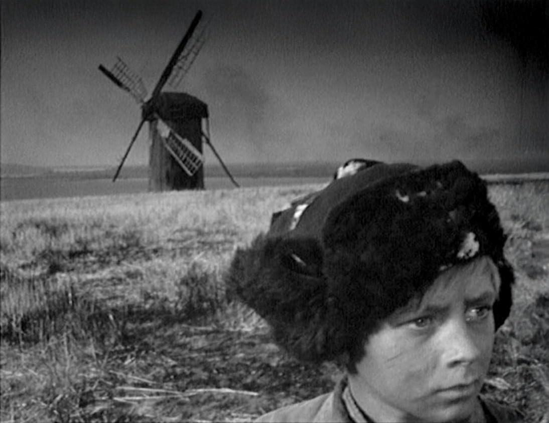 Ein Junge mit Pelzmütze blickt nach rechts. Im Hintergrund steht eine Windmühle auf einem grasbewachsenen Hügel. Der Himmel ist bedeckt.