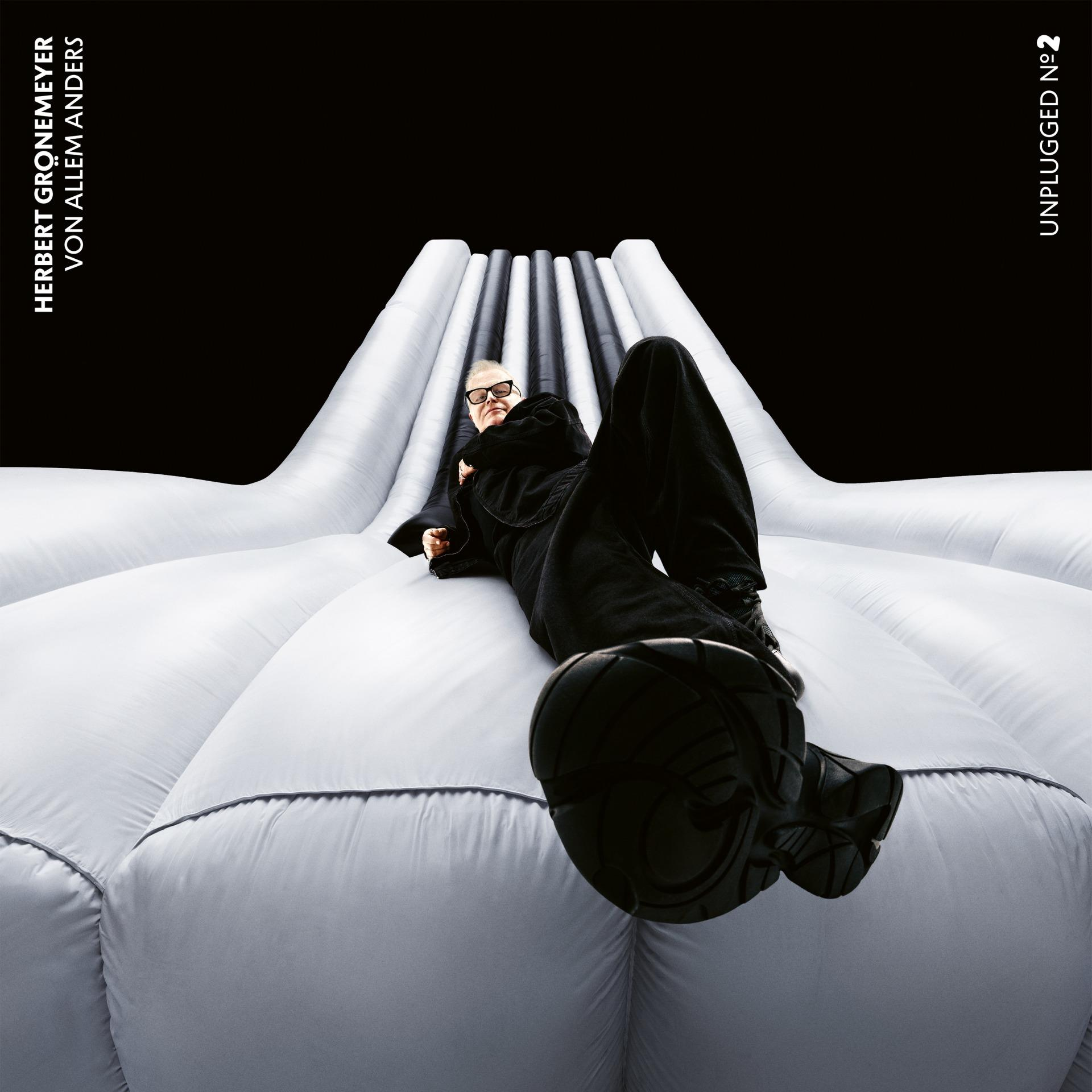 Mann in schwarzem Anzug und Brille liegt auf weißer aufblasbarer Struktur vor schwarzem Hintergrund. Albumcover.