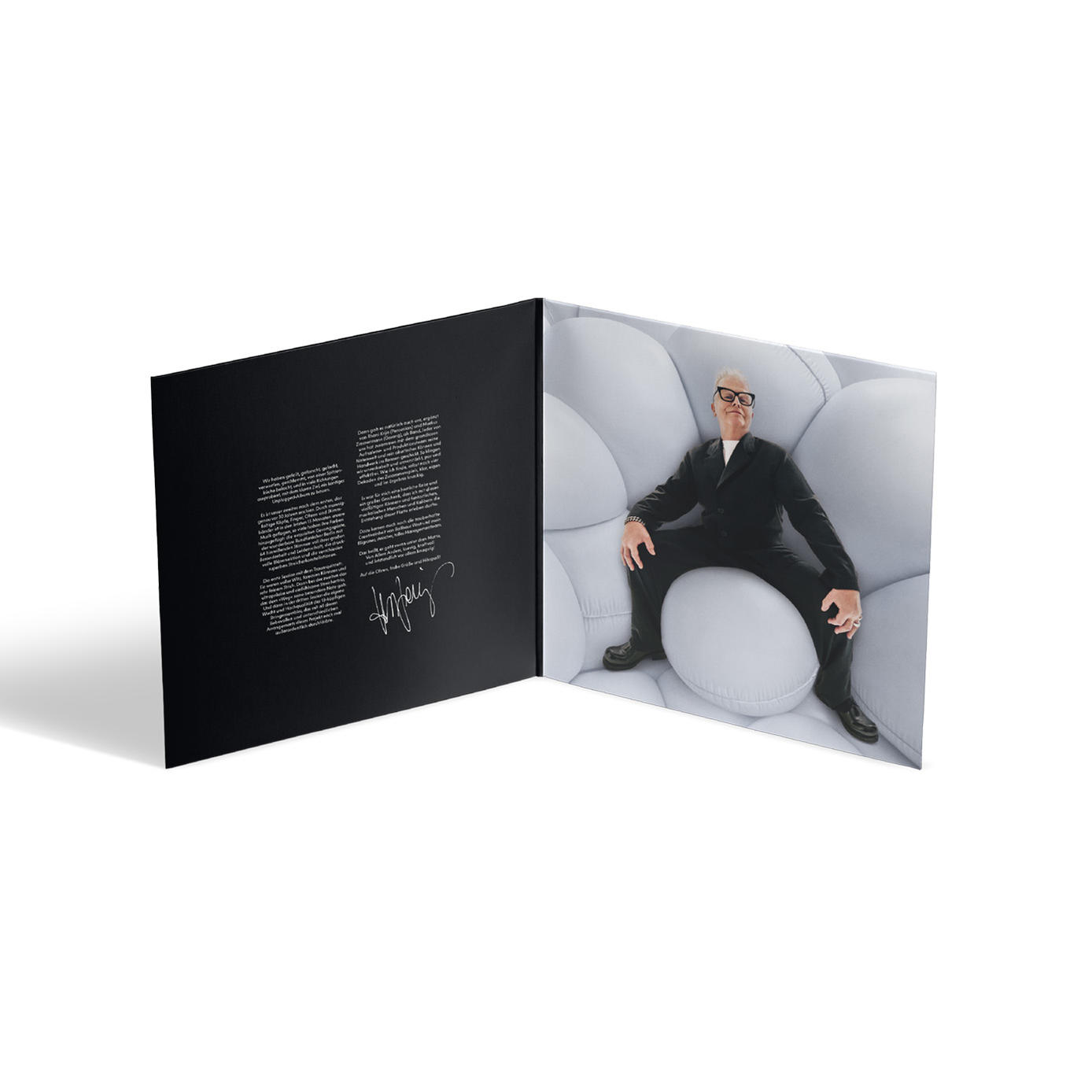 Geöffnetes Albumcover, Mann in schwarzem Anzug, sitzt auf großem weißen Kissen. Linke Seite ist schwarzer Text mit Unterschrift. Weißer Hintergrund.