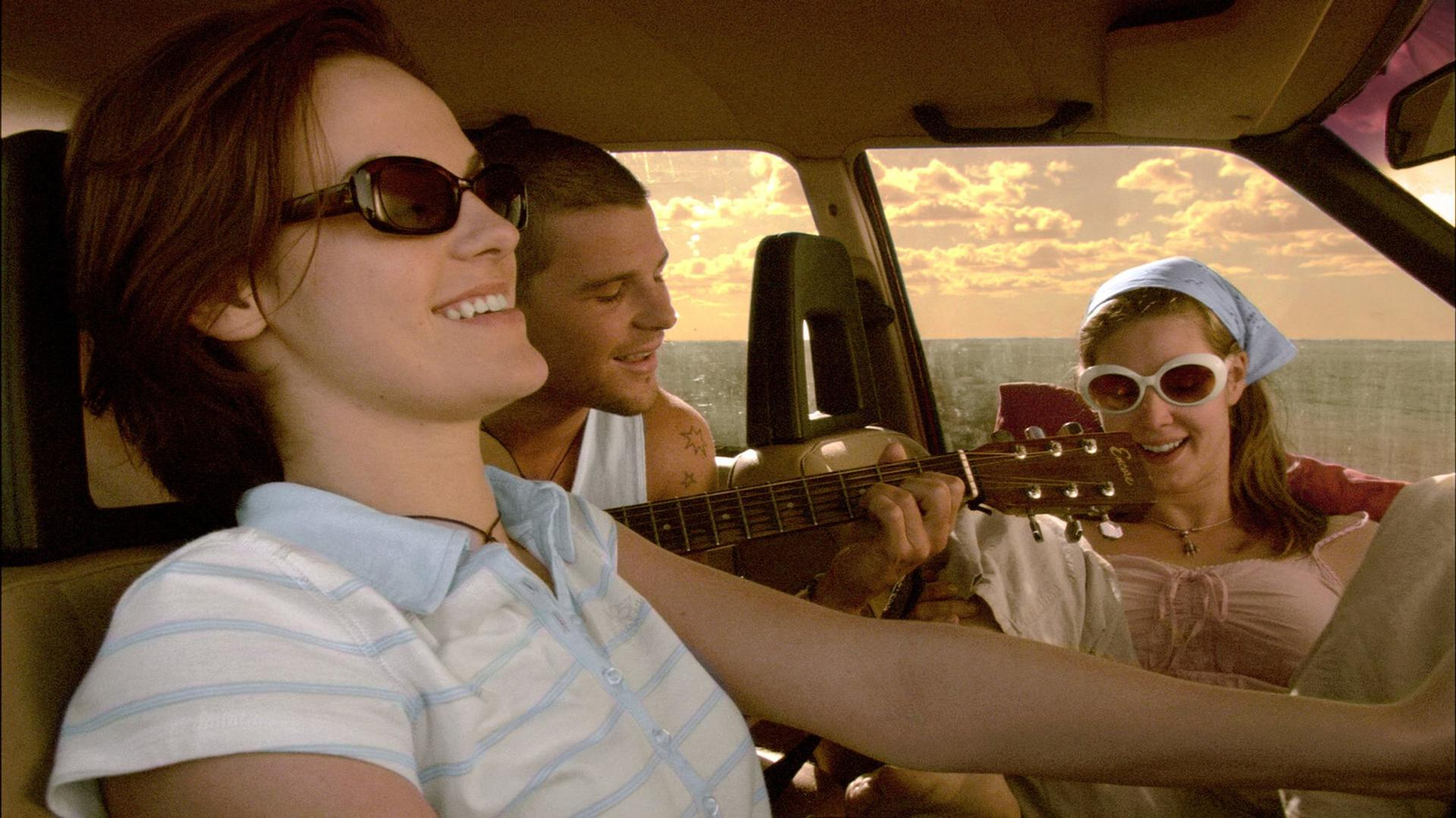 In einem Auto lächeln drei Personen. Einer trägt eine Sonnenbrille und ein gestreiftes Hemd. Ein anderer spielt Gitarre. Himmel sichtbar.