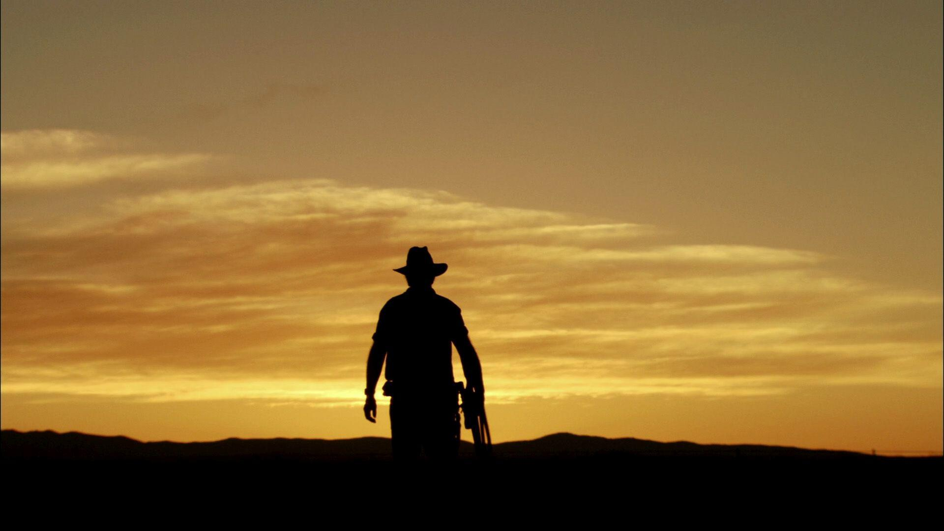 Silhouette eines Cowboys mit Hut und Gewehr, der auf den Sonnenuntergang zugeht. Oranger Himmel mit Wolken, dunkle Landschaft im Hintergrund.