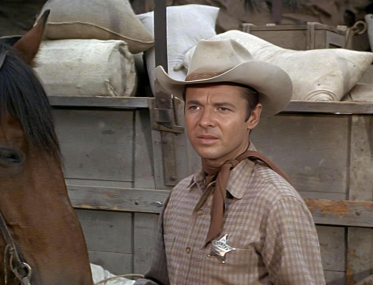 Ein Mann mit Cowboyhut und Sheriffabzeichen steht in der Nähe eines Pferdes. Der Hintergrund umfasst Säcke und Holzkonstruktionen.