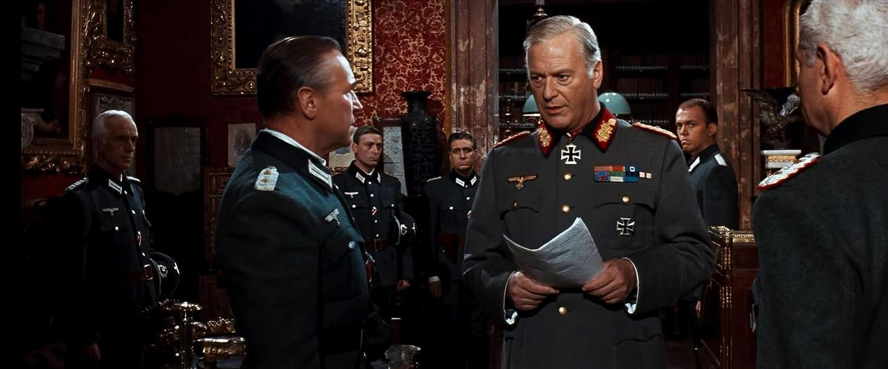 Mehrere Männer in Militäruniformen versammeln sich in einem Raum. Einer hält Papiere. Der Raum hat dunkle Holzmöbel und rote Tapeten.