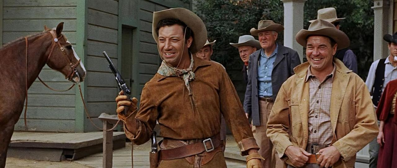 Eine Gruppe Cowboys in Westernkleidung. Einer hält eine Waffe und lächelt. Ein braunes Pferd steht daneben. Andere schauen zu, lachen und lächeln.