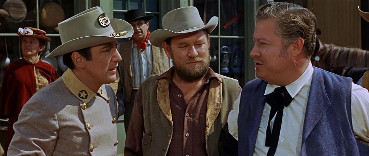 Drei Männer mit Cowboyhüten unterhalten sich vor einem Geschäft. Andere im Hintergrund. Die Szene spielt im Wilden Westen.