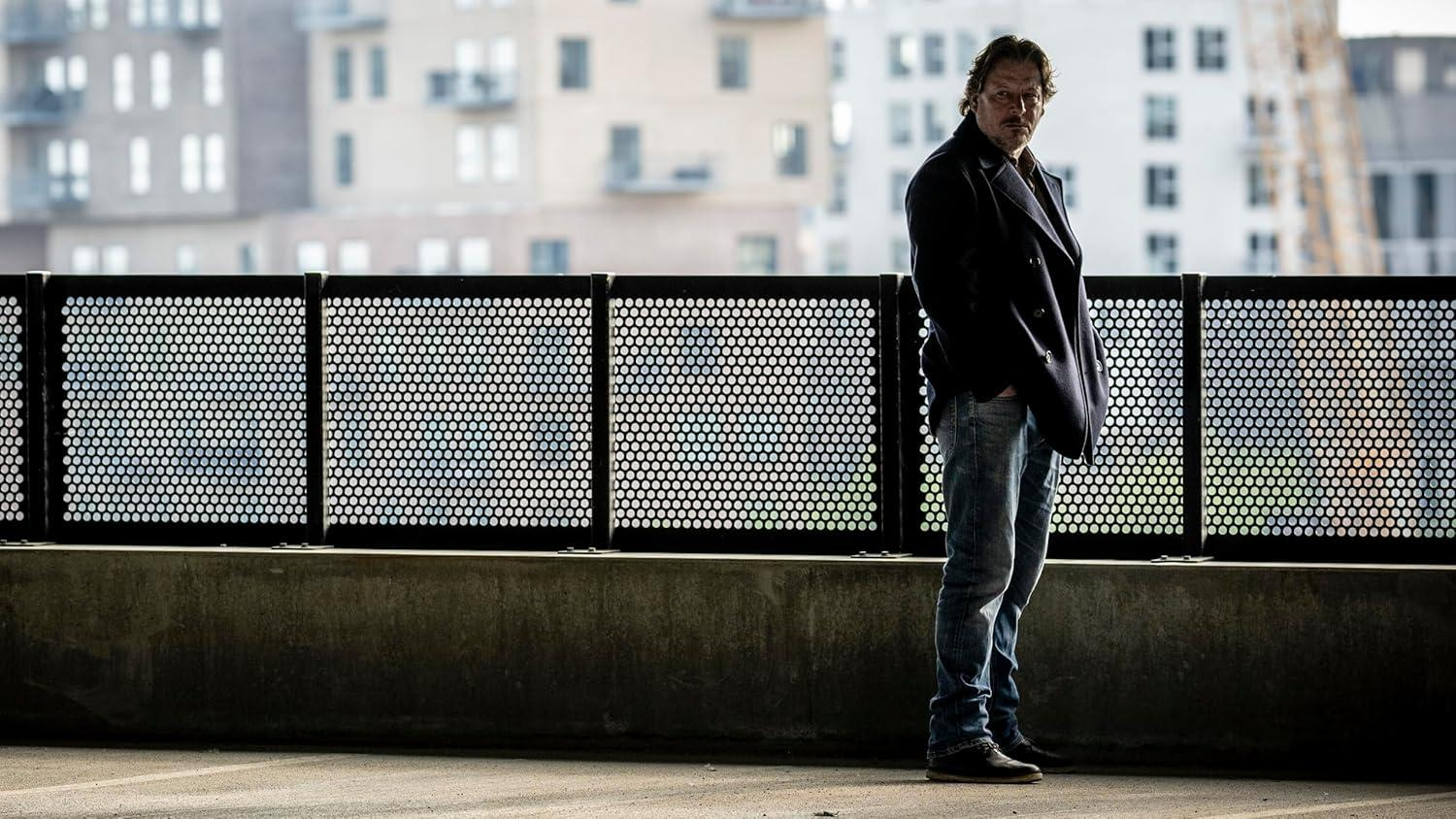 Ein Mann in einem dunklen Mantel und Jeans steht in der Nähe eines Metallzauns, im Hintergrund Gebäude. Der Mann schaut in die Kamera.