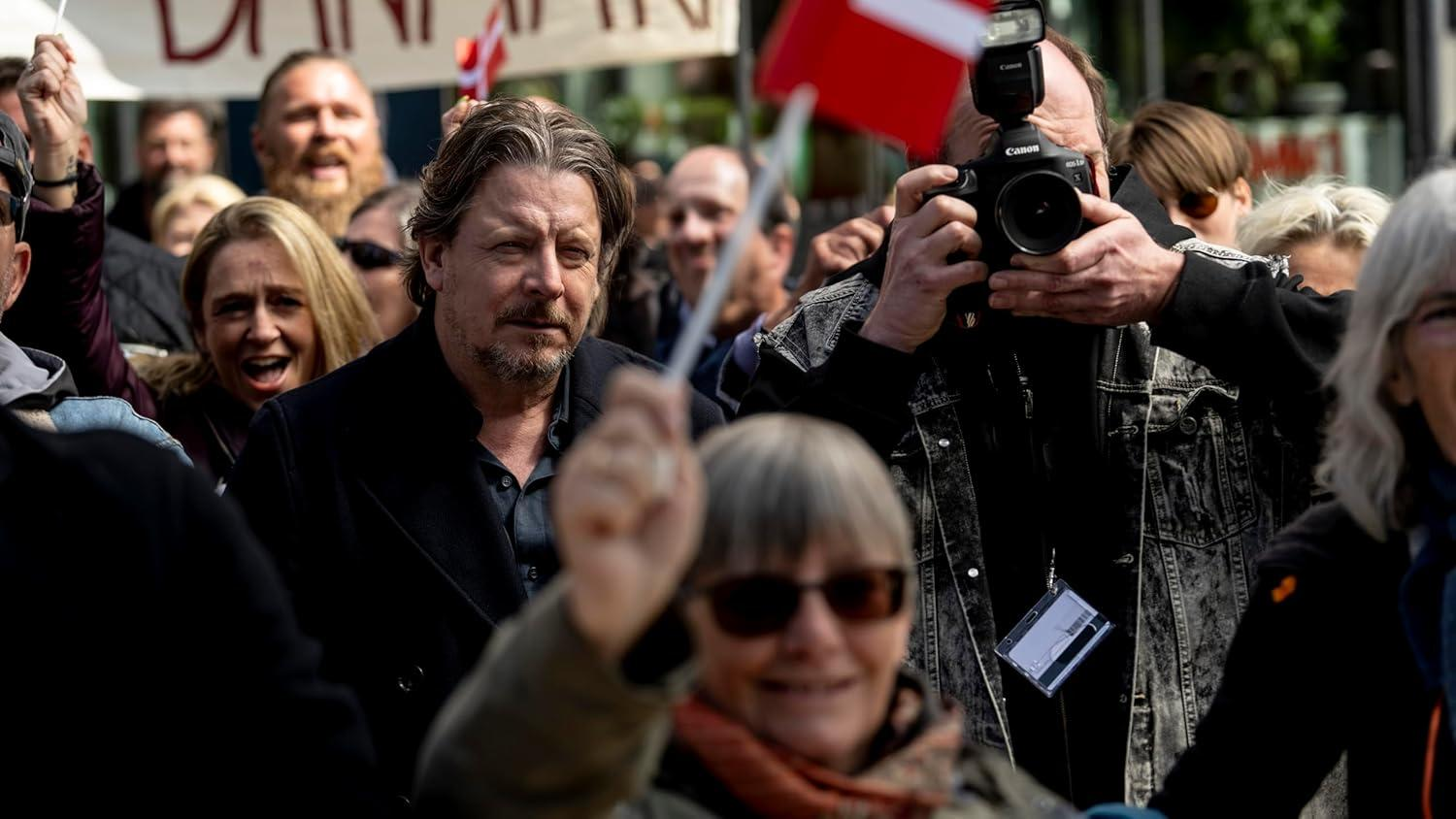 Menschen bei einer Kundgebung mit Fahnen und Bannern. Ein Mann mit Kamera macht ein Foto. Andere lächeln und jubeln.