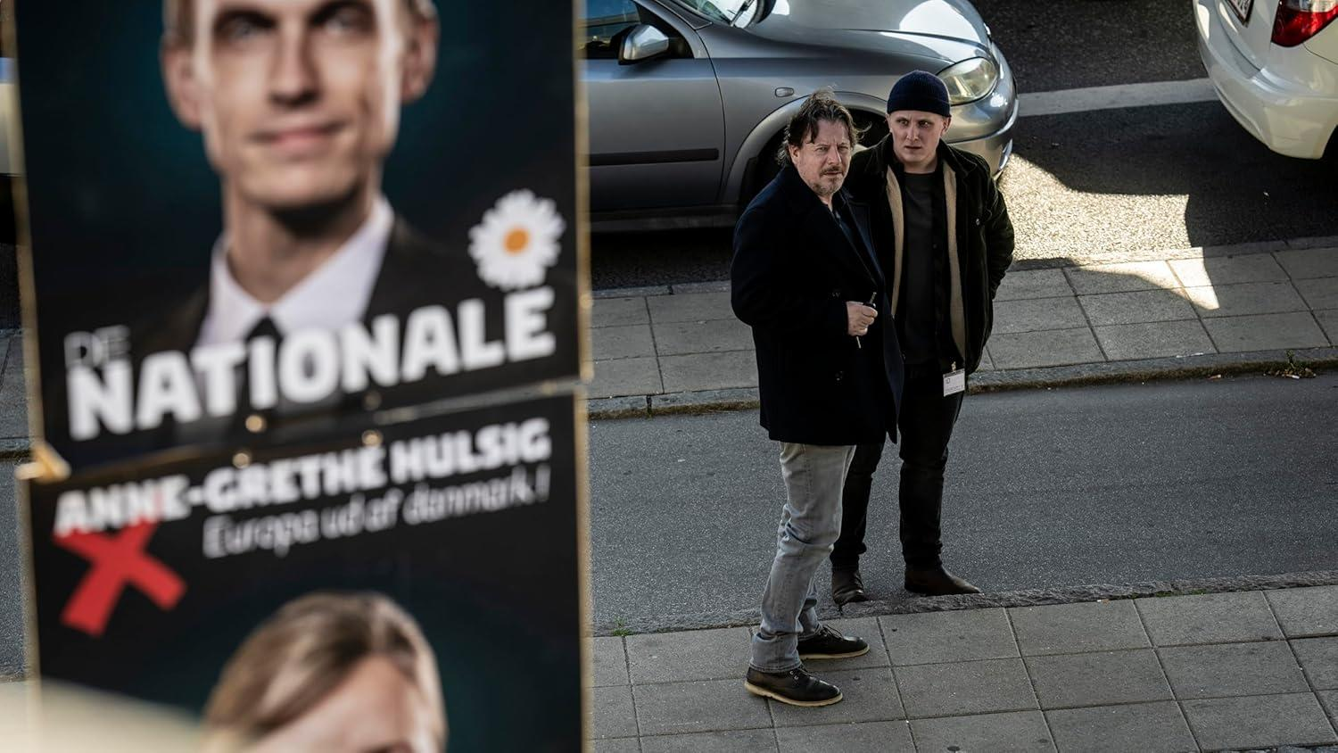 Zwei Männer stehen in der Nähe eines politischen Plakats. Das Plakat zeigt das Gesicht eines Mannes und dänischen Text. Autos sind hinter ihnen geparkt.