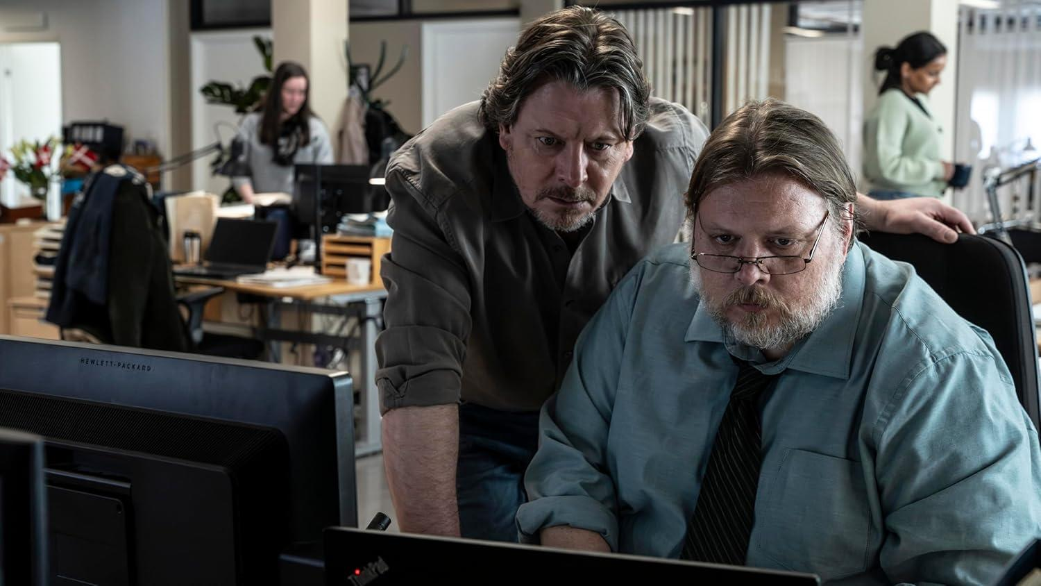 Zwei Männer betrachten einen Computerbildschirm in einem Büro. Andere arbeiten im Hintergrund. Ein Mann trägt eine Brille und eine Krawatte.