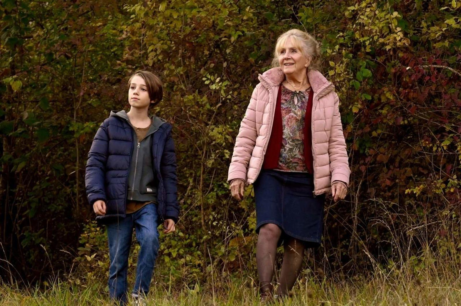 Ein Junge und eine ältere Frau stehen auf einem Feld mit einem Wald im Hintergrund. Die Frau trägt eine rosa Jacke, der Junge eine blaue.