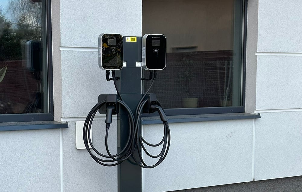 Két elektromosautó-töltő egy szürke oszlopon, a kábelek lógnak. Épület ablakokkal a háttérben. Visszaverődéses üveg és világosszürke falak.