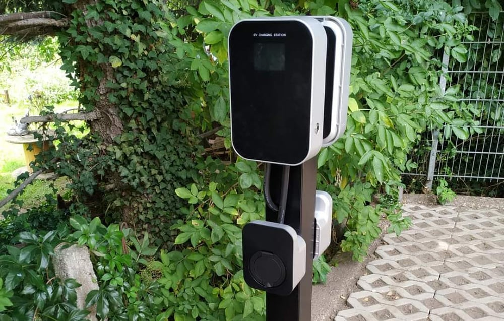 Egy fekete képernyős és dugós EV töltőállomás zöld lombozat előtt. Balra egy borostyánnal borított fa látható.