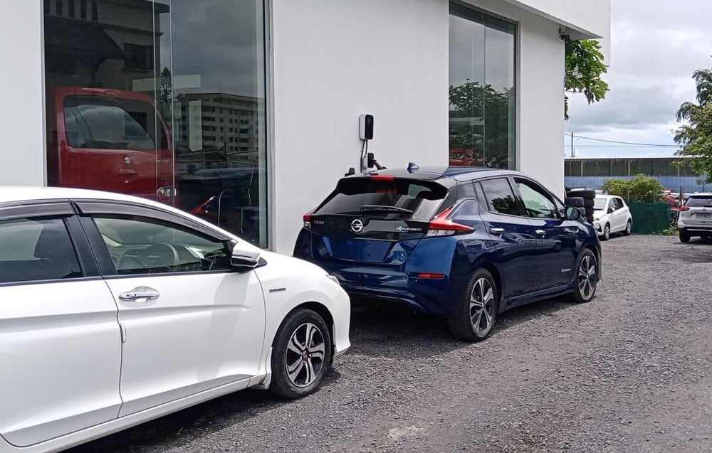 Fehér autó és kék elektromos autó parkol egy üvegablakos épület közelében. Piros autó látható a tükröződésben.