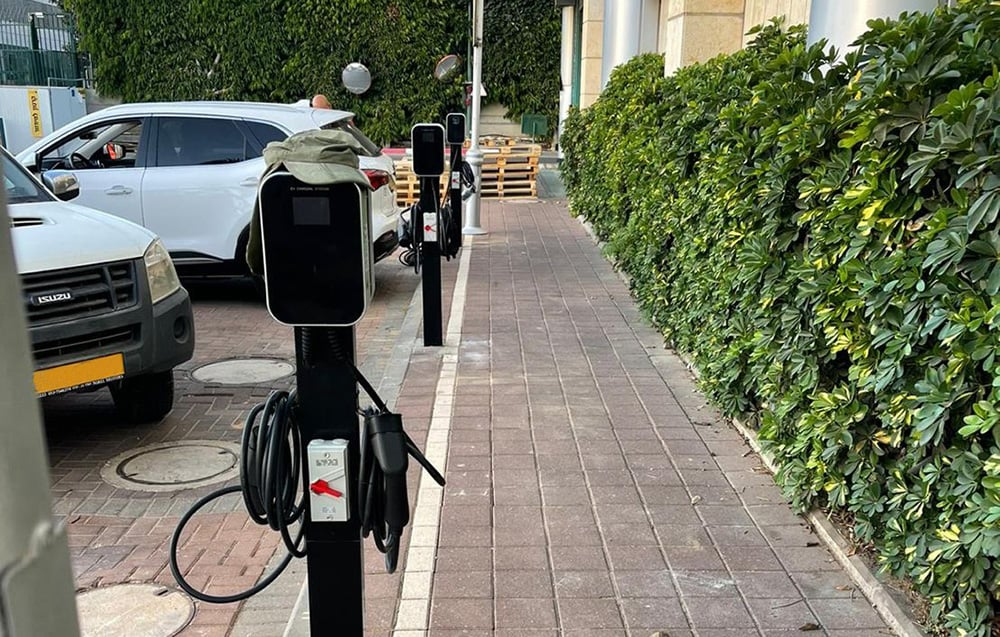 Elektromos autó töltőállomások egy sövény mellett. Egy fehér SUV és egy fehér teherautó parkol a közelben. Egy téglajárda van jobbra.