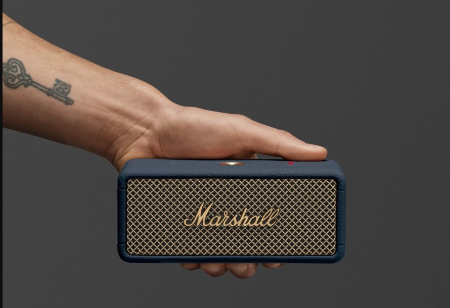 Eine Hand hält einen Marshall-Lautsprecher. Der Lautsprecher ist marineblau mit goldfarbener Gitterfront. Ein Schlüssel-Tattoo ist auf dem Arm.