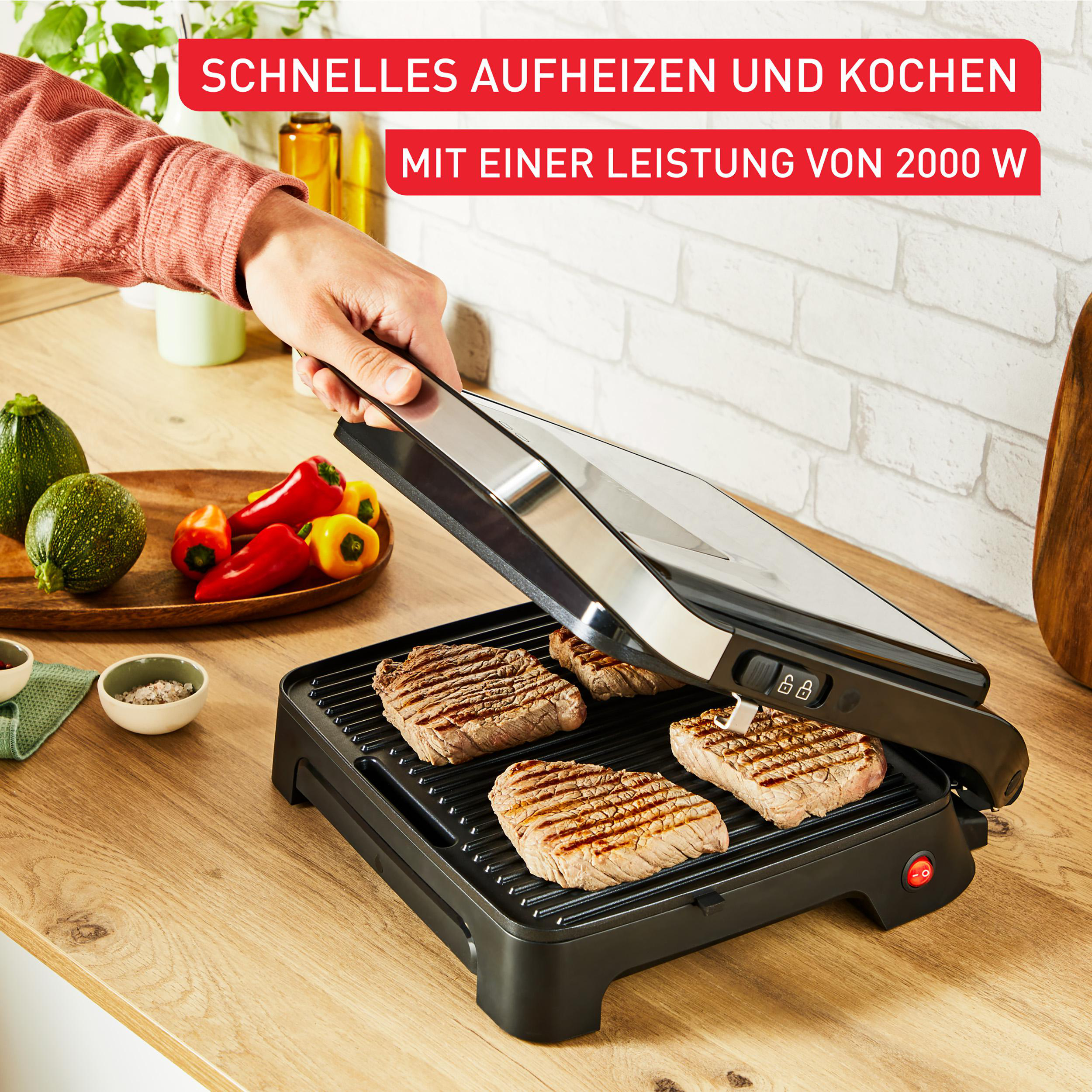 Eine Person grillt Steaks mit einem schwarzen Grill, Gemüse daneben. Im Hintergrund eine weiße Ziegelmauer.