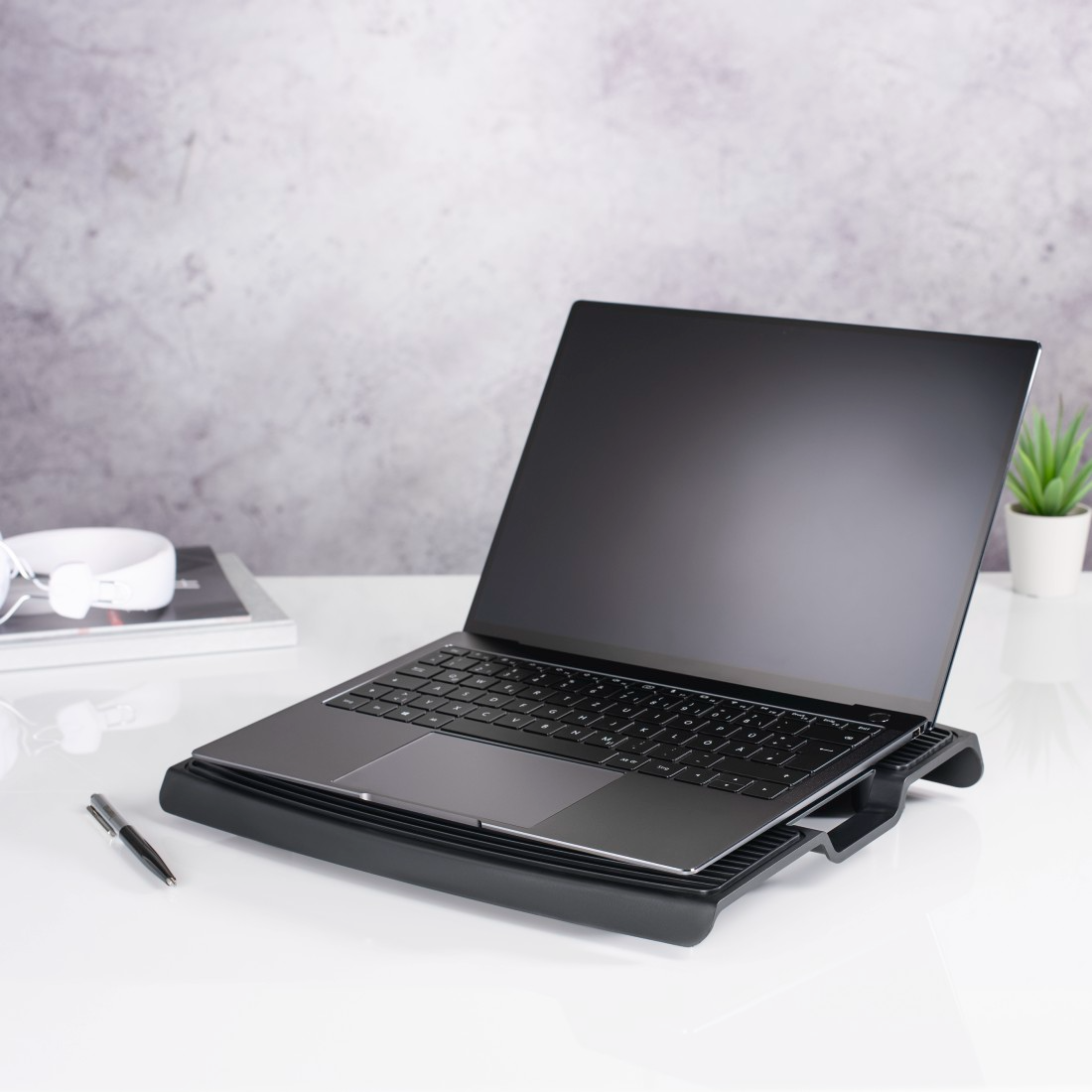 Egy laptop egy fekete hűtőpárnán egy fehér asztalon. Fejhallgató és egy növény a háttérben. Egy toll az asztalon.