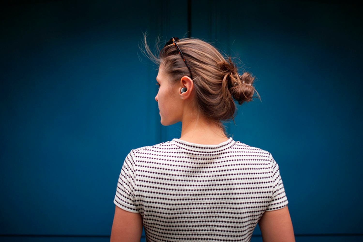 Mujer con moño y gafas en la cabeza con una camisa a rayas. Tiene un auricular en la oreja, frente a una pared azul.
