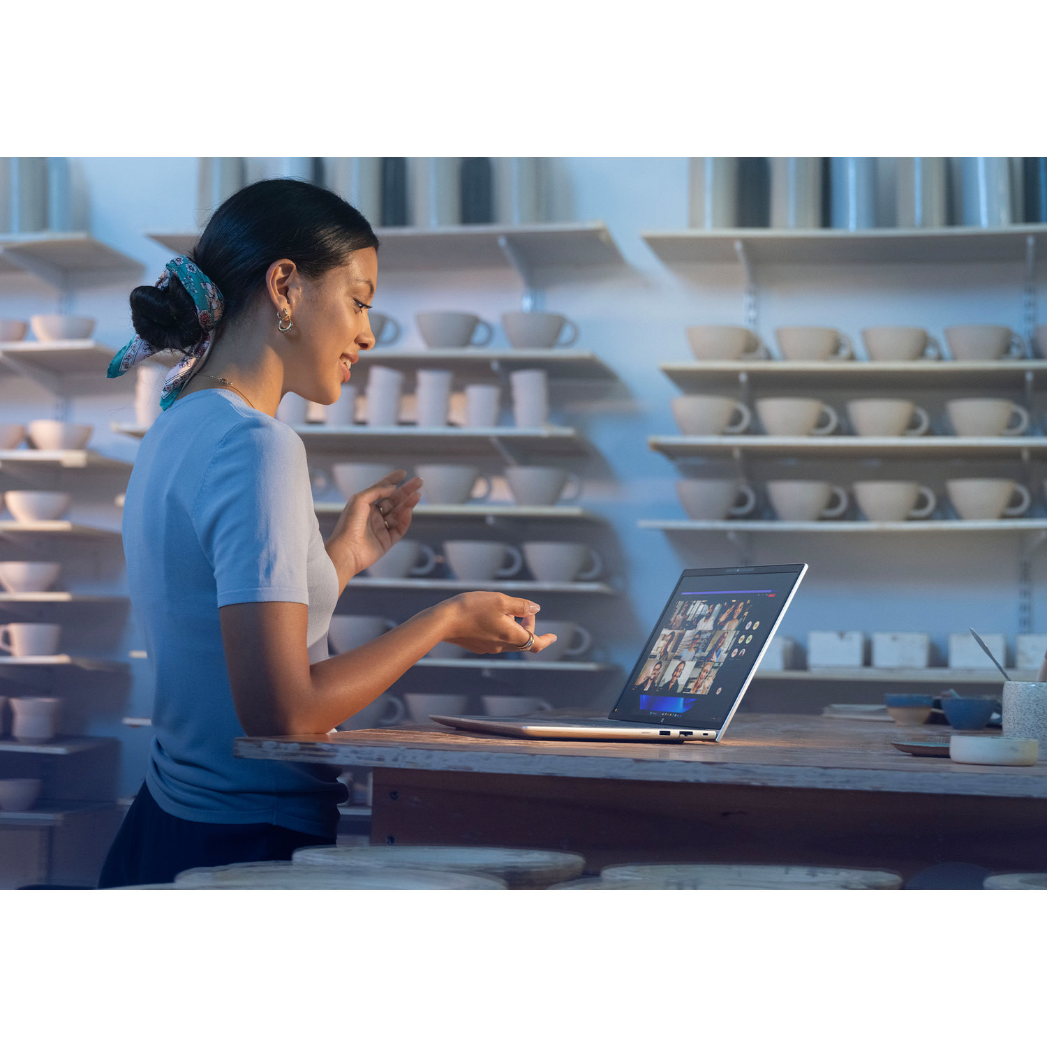 A woman in a pottery studio on a video call, gesturing. Shelves of pottery line the background. Laptop sits on a wooden table.
