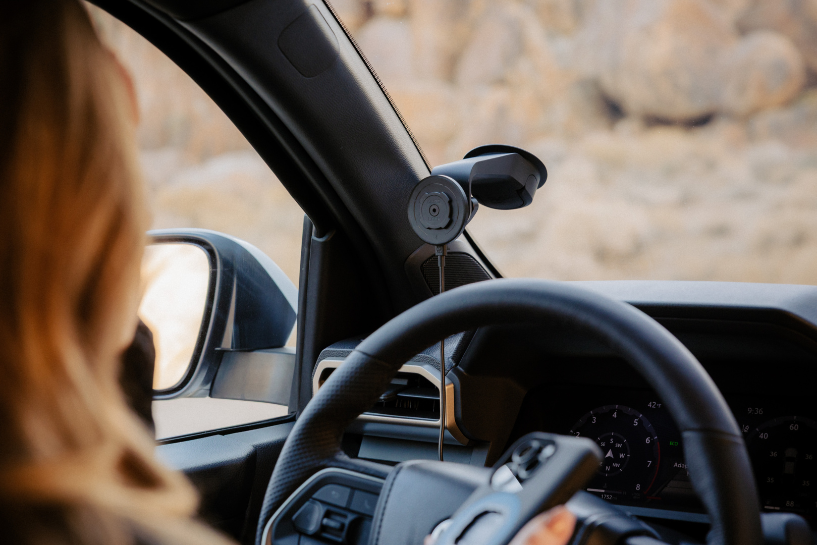 All'interno di un'auto, sono visibili volante e cruscotto. Sono presenti un supporto per telefono e uno specchietto retrovisore. Una persona sta guidando.