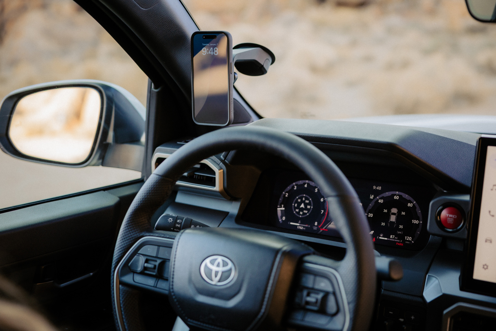 Interni auto con cruscotto, volante, telefono e specchietto visibili. Il telefono mostra le 9:48 e la data. Sfondo paesaggistico.