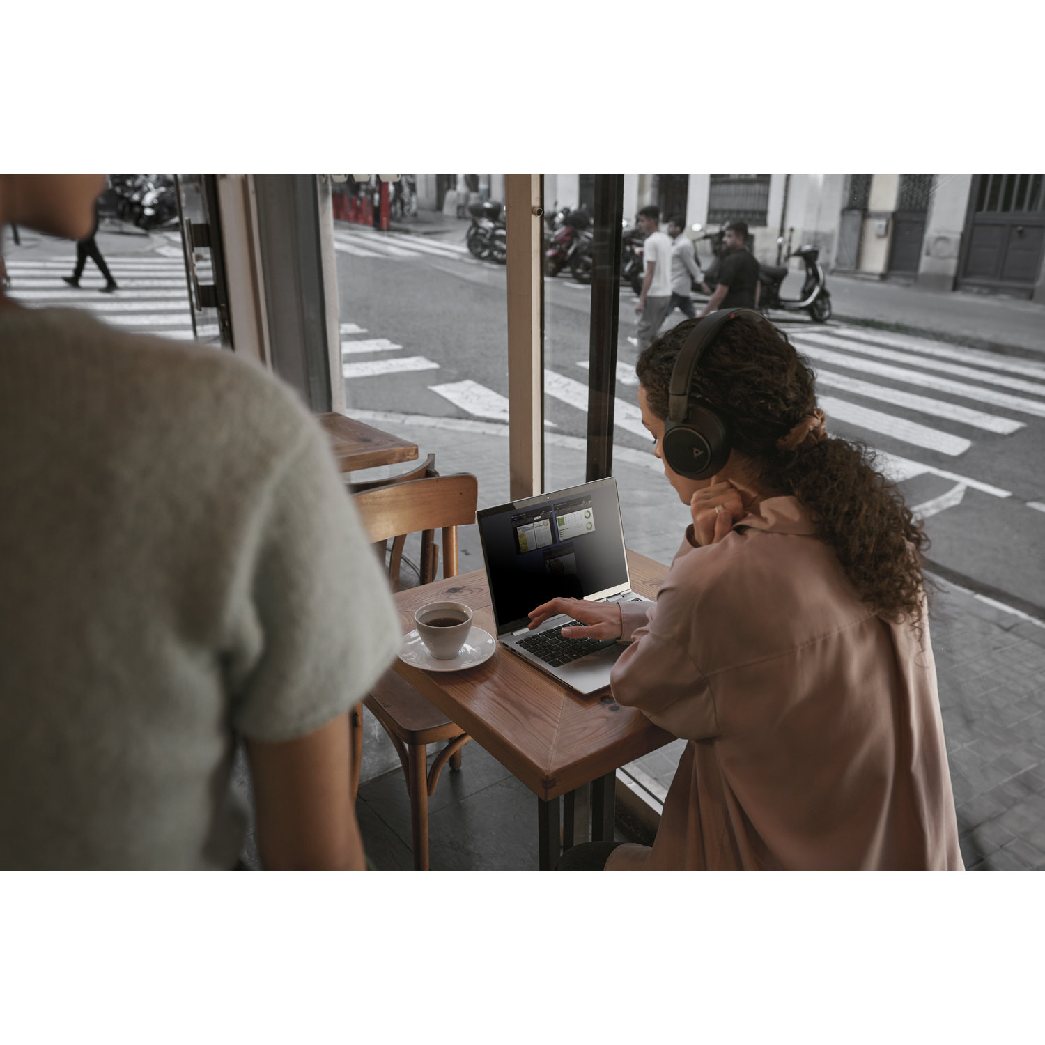 Woman wearing headphones works on laptop at cafe. A coffee cup sits on the table. People walk outside the window.