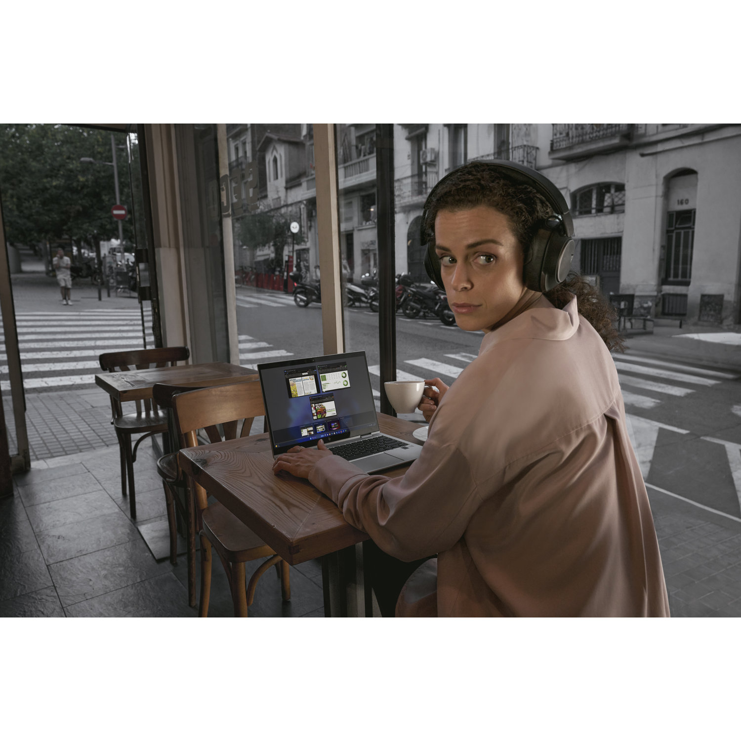 Woman wearing headphones in a cafe, using a laptop. She looks back towards the viewer and holds a mug, with street scene in the background.