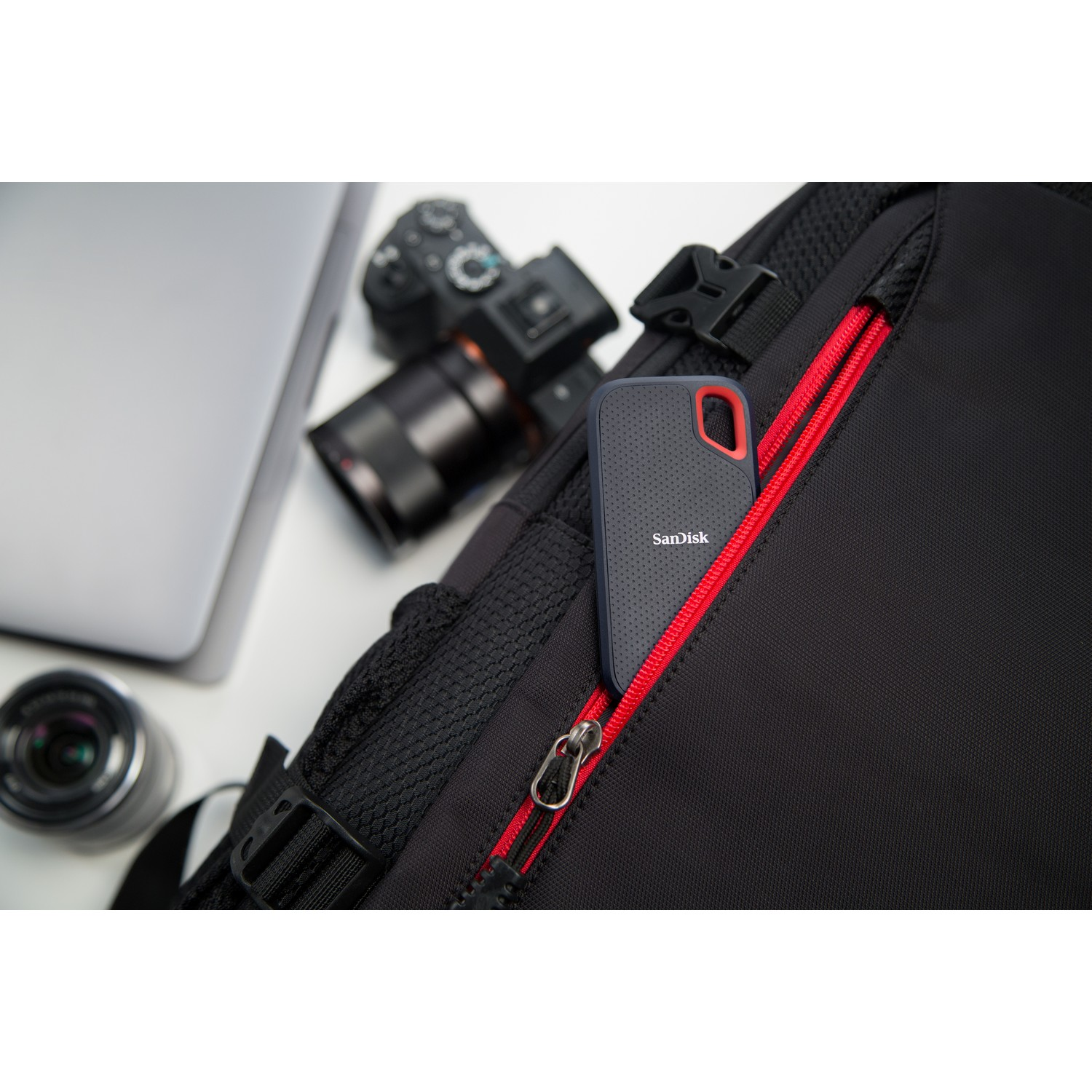 Una mochila negra con cremallera roja contiene un SSD SanDisk. Un portátil y una lente de cámara están sobre una superficie blanca.
