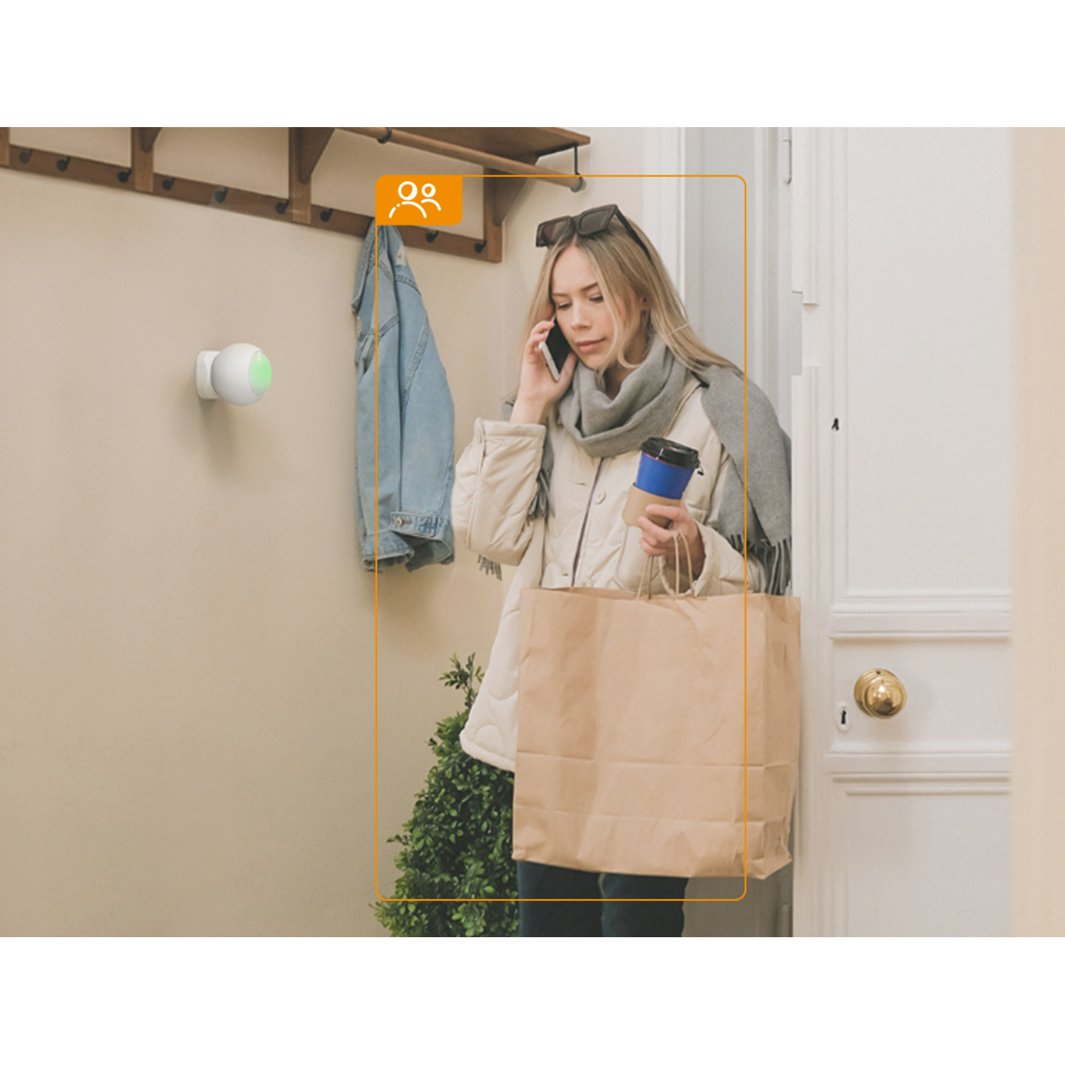 A woman is on the phone holding a coffee cup and a paper bag. A camera on the wall, a coat, and a door are also visible.