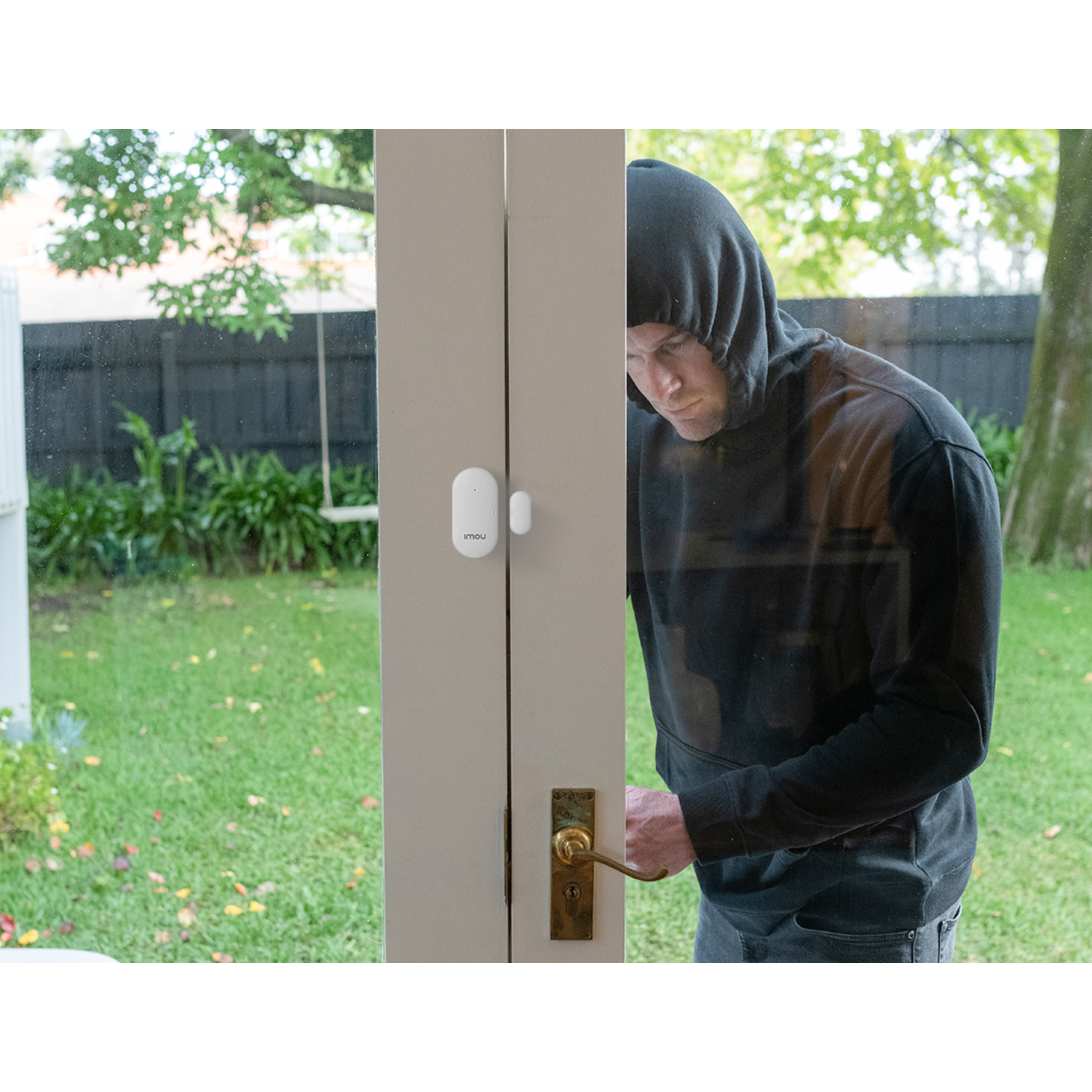 A man in a hoodie is attempting to open a door, likely breaking in. A security sensor is on the door. Green lawn in background.