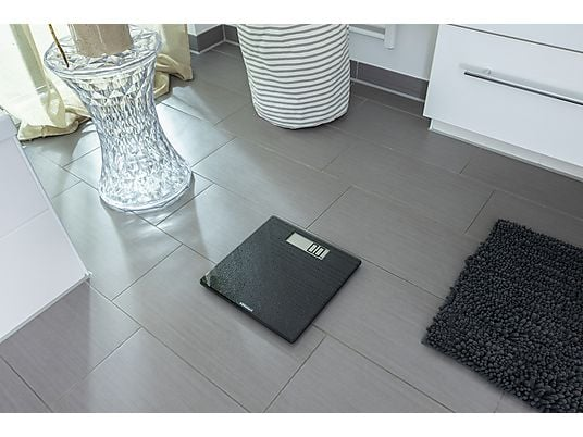 Escena de baño con báscula negra, baldosas grises, alfombra pequeña, taburete decorativo y cesta de lavandería a rayas.