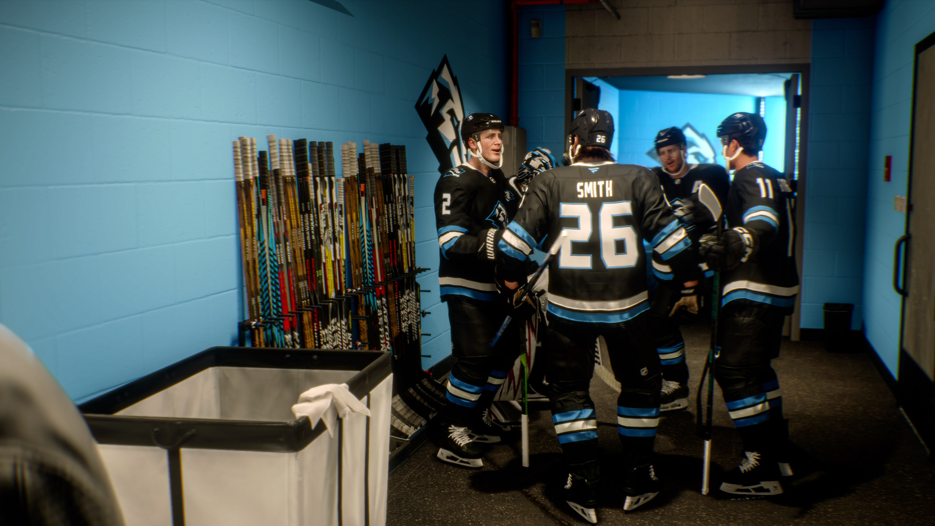 Eishockeyspieler in einer Umkleidekabine, tragen schwarz-blaue Trikots. Hockeyschläger lehnen an einer blauen Wand. Eine Wäschetonne ist im Vordergrund.