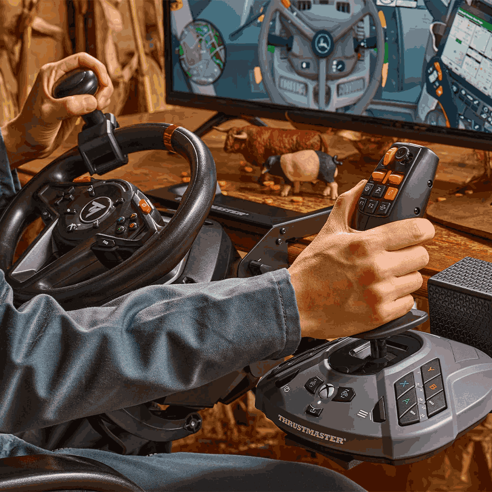 Manos en una configuración de volante y joystick para juegos. Un monitor muestra un juego de granja con ganado. La configuración está sobre una superficie de madera.