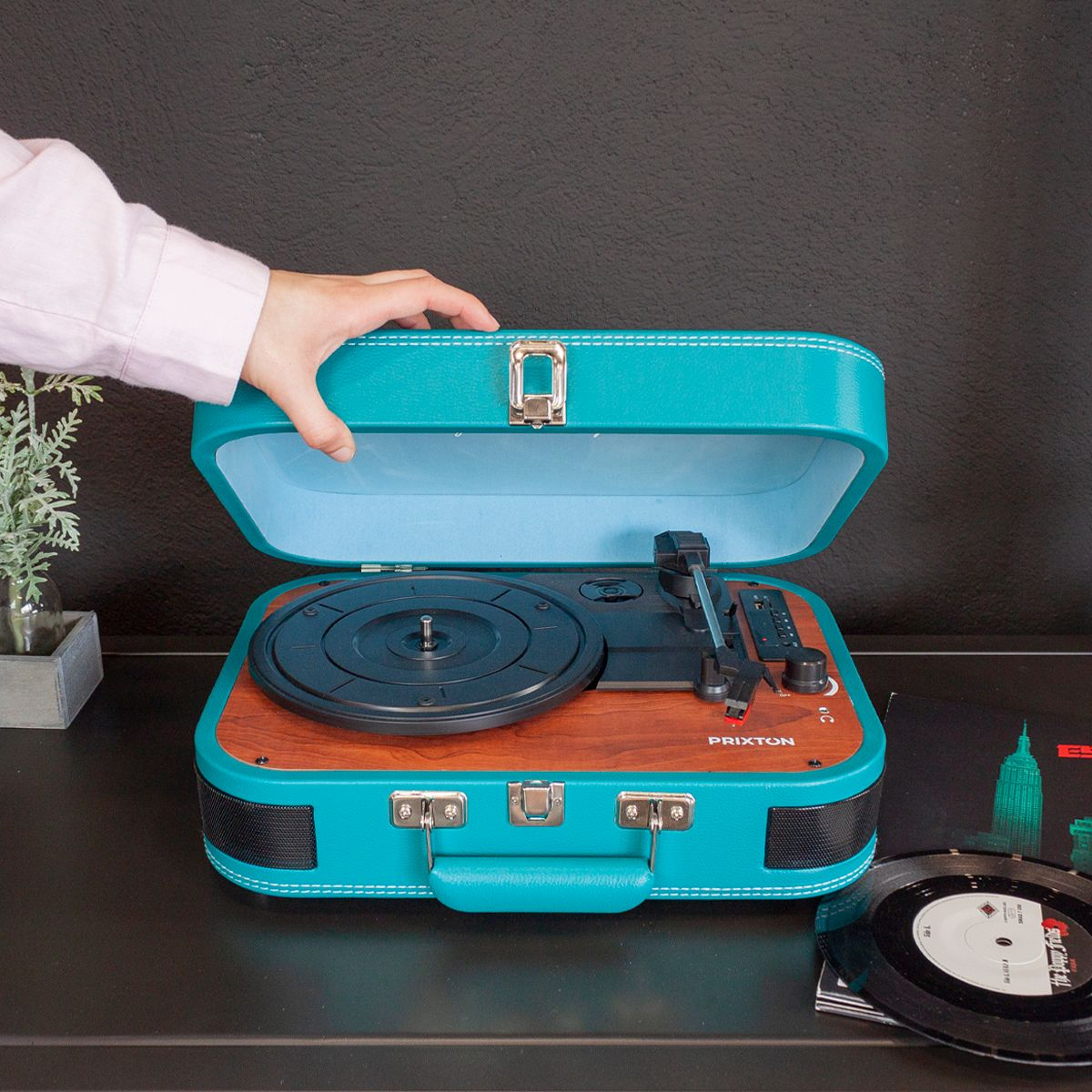 Un tocadiscos turquesa con la tapa abierta. Una mano lo está abriendo. Un disco y una pequeña planta están cerca. El fondo es negro.