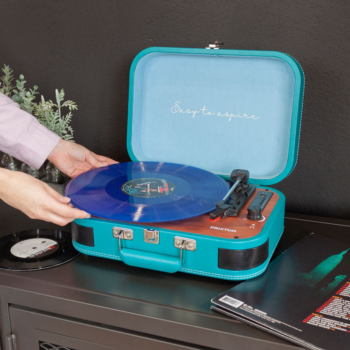Manos colocando un disco de vinilo azul en un tocadiscos turquesa tipo maleta. Un disco negro está en una mesa junto a él.