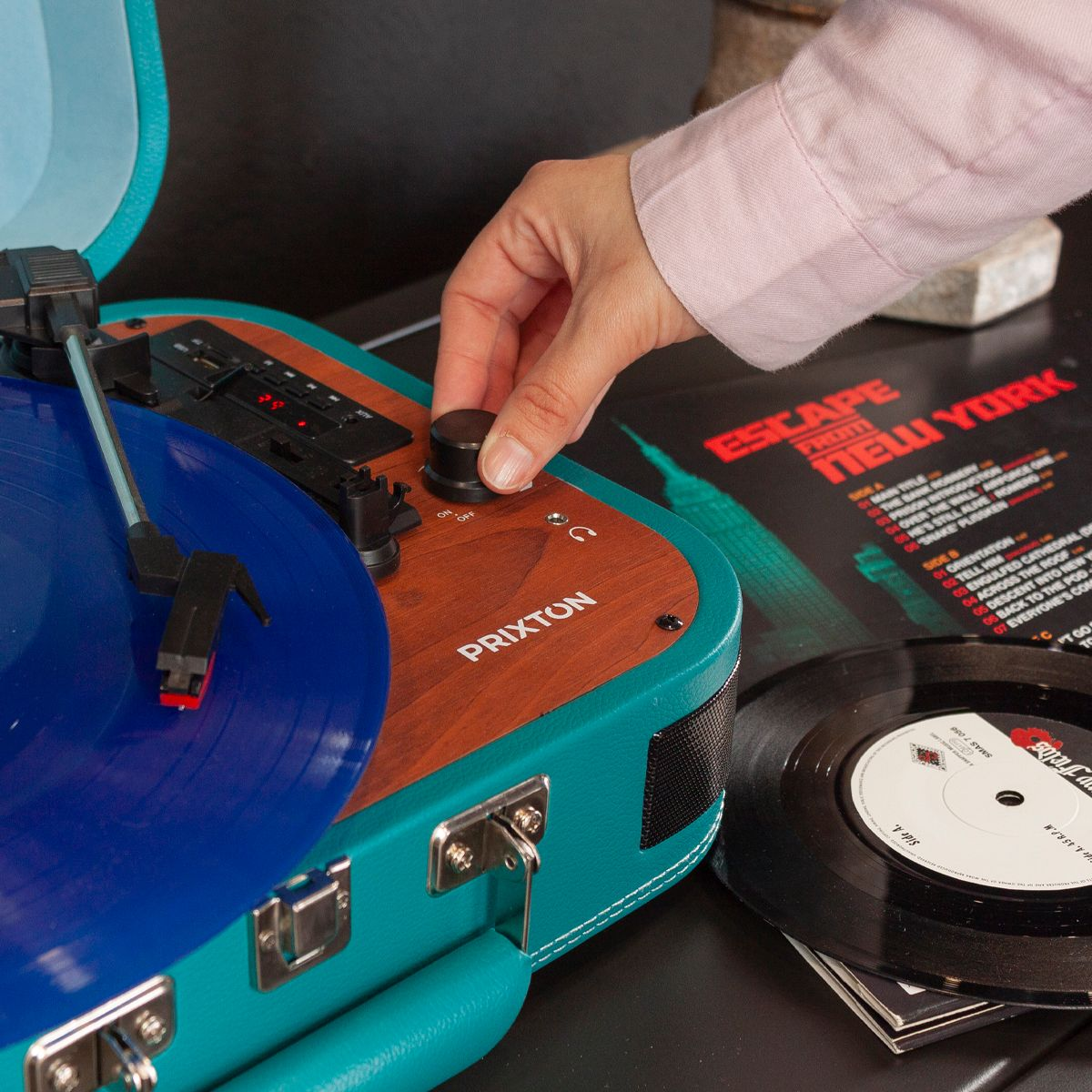 Una persona ajusta la perilla de volumen de un tocadiscos. Un disco de vinilo azul está en el reproductor. Una portada de álbum es visible en el fondo.